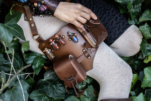 Close-up of the brown leather leg holster worn on a LARP costume, with colorful glass potion vials in handmade holders and a hand opening the upper pouch. Crafted by Outcast Props from full-grain vegetable-tanned leather with brass rivets, it combines utility and fantasy style. Perfect for rogues and alchemists in cosplay, renaissance fairs, or immersive medieval adventures seeking authentic handcrafted design.
