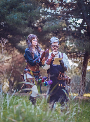 Two LARP players in a forest wearing Outcast Props leather gear, including the rogue leg holster with colorful potion vials and matching pouches. Both costumes feature belts, scrolls, and accessories inspired by fantasy alchemists. Made from vegetable-tanned leather, this potion set adds realism, color, and authentic medieval flair to cosplay, roleplay, and renaissance fair outfits with detailed artisan quality.