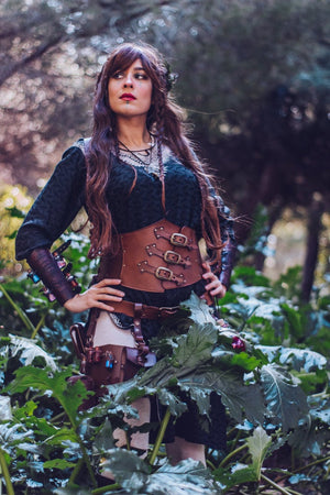 Female LARP character standing among green leaves, wearing the brown leather rogue leg holster by Outcast Props along with a corset belt and potion vials. Handcrafted from vegetable-tanned leather with brass buckles and embossed detail, this fantasy accessory enhances medieval costumes for cosplay, renaissance fairs, or alchemist characters. Combines elegance, strength, and practicality for immersive adventures.