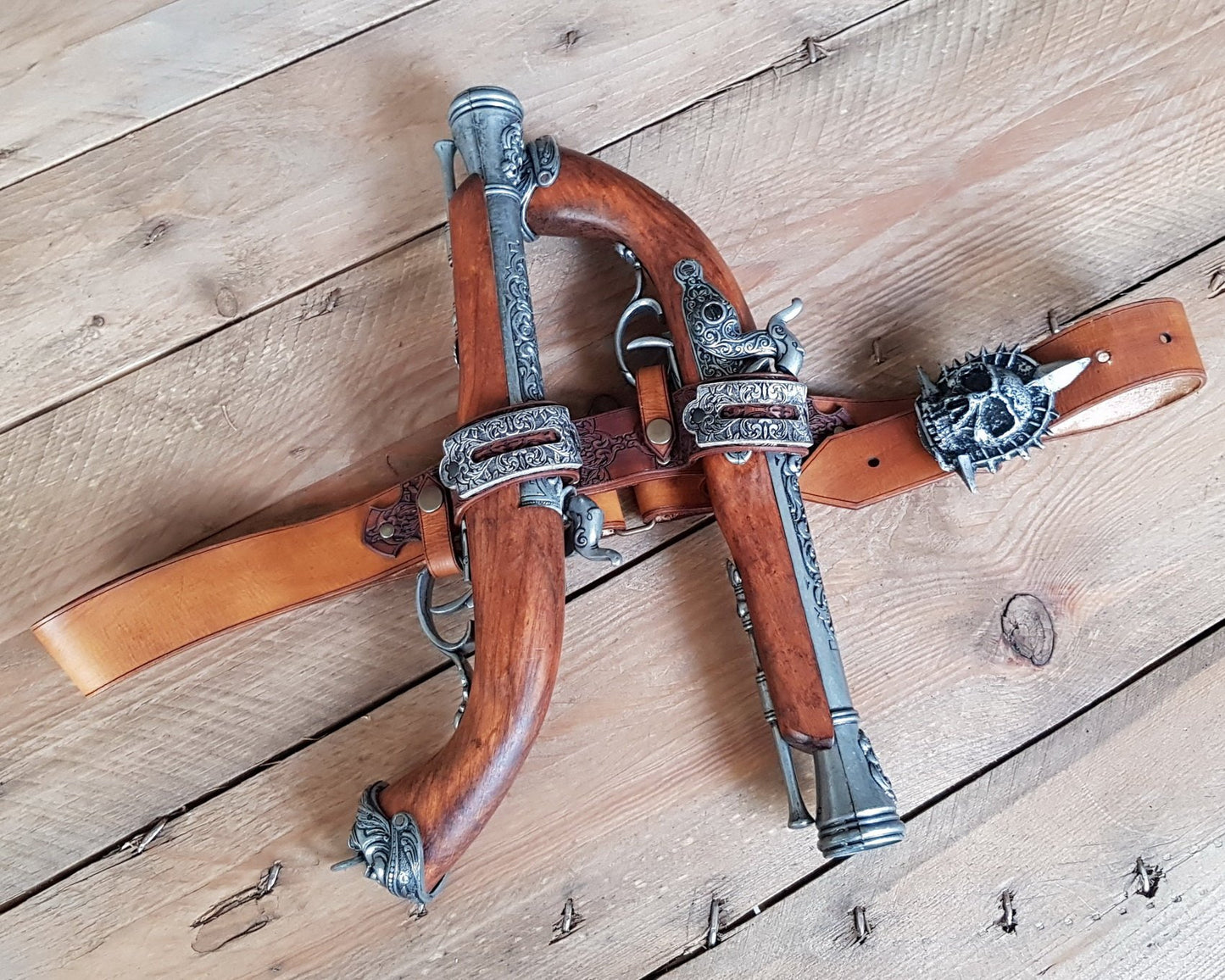 Tan leather pirate baldric arranged with two flintlock pistols, engraved holsters, and a spiked skull emblem on rustic wood. Styled after Edward Kenway from Assassins Creed Black Flag, this accessory is ideal for pirate cosplay, LARP, or steampunk rogue outfits. Authentic feel, handcrafted for epic adventures and bold character builds.