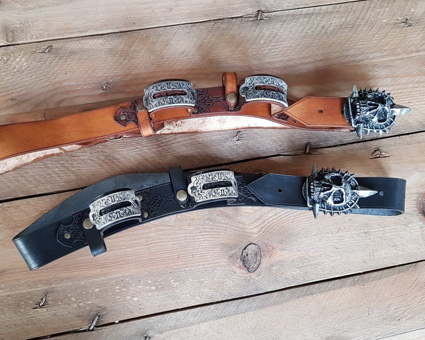 Two detailed pirate baldrics laid flat on wooden planks—one in black leather, one in tan. Each baldric features antique silver buckles and a spiked skull emblem inspired by Edward Kenway and Assassins Creed Black Flag. These leather gun holsters are perfect for pirate cosplay, steampunk adventurers, or historical reenactment. Crafted for quality and immersive roleplay.