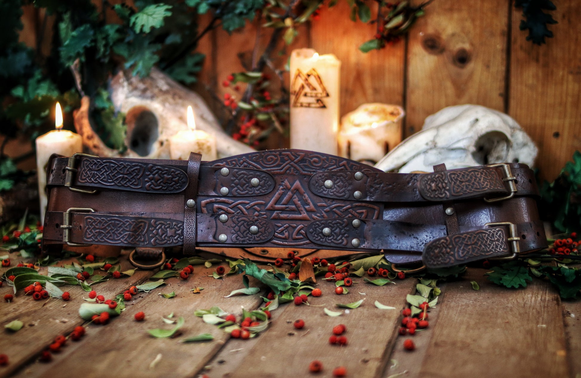 Valknut viking belt placed between burning candles and animal skulls on a wooden floor with leaves and berries. Emphasizes the mystical LARP aesthetic. Long tails: viking leather armor belt, celtic fantasy belt, renfaire costume wide, barbarian cosplay outfit, fantasy medieval warrior accessory. Perfect for immersive roleplay photoshoots.