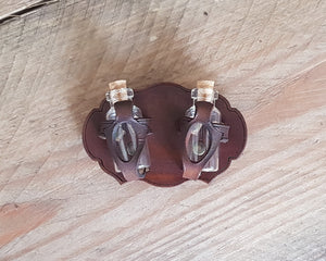 Two small potion bottles attached to a brown leather holder with a hollow space for viewing the bottle contents, placed on a wooden surface. Belt Potion holder for two small bottles. Made of leather for adventurers, steampunk, alchemist, healers, cosplay or just to keep spices