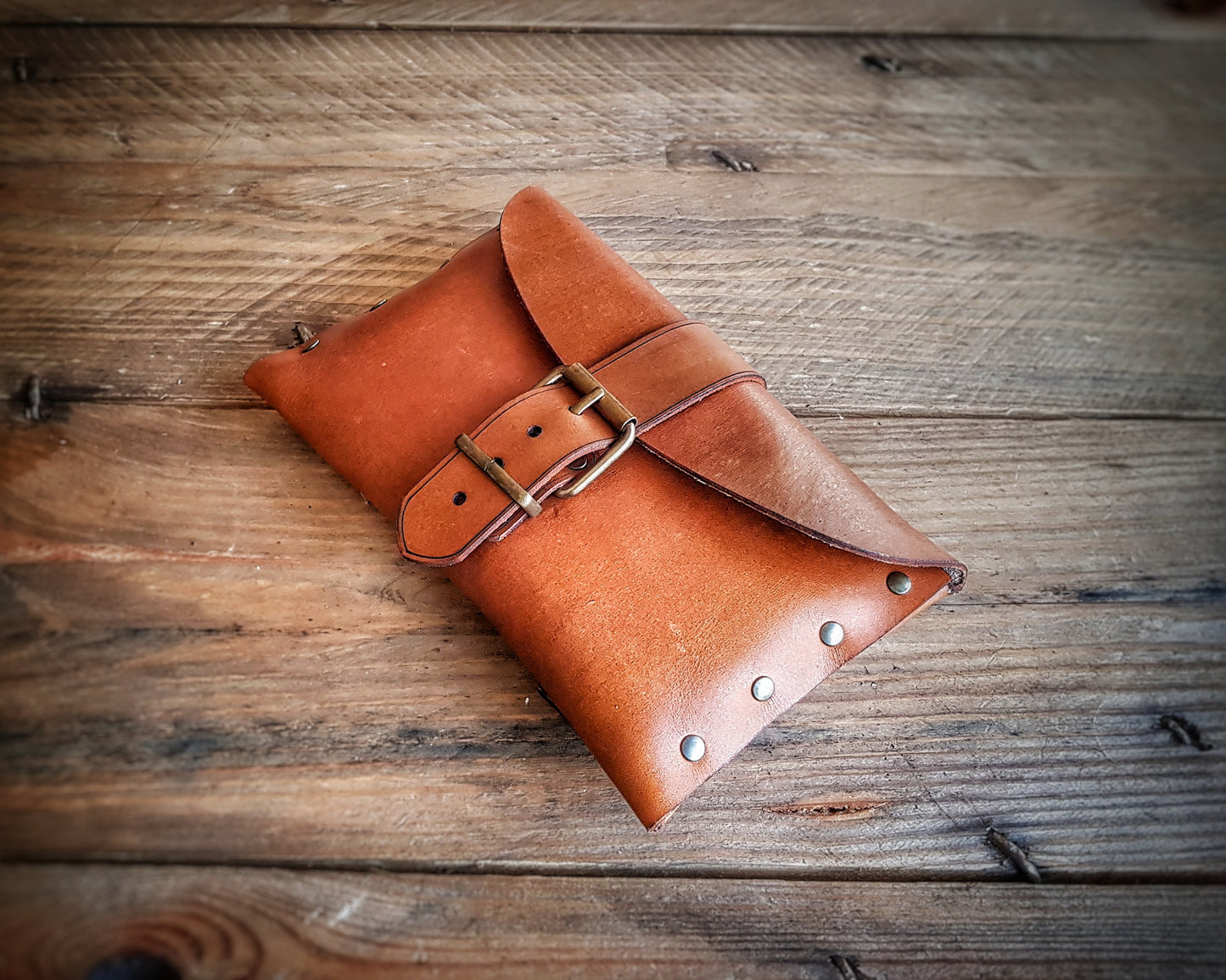 Medieval leather pouch with buckle closure placed on rustic wooden surface. Handmade Renaissance fair leather bag with natural brown tone, brass studs and curved flap. Perfect for LARP costume accessory or fantasy belt bag. Shows artisan texture of vegetable-tanned leather. Ideal for viewers needing a detailed tactile and visual description.