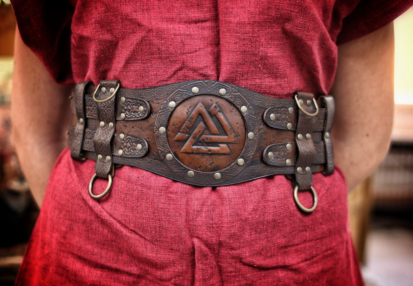Brown viking leather armor belt on female model in red dress with fur shoulder cape. The belt shows Norse knotwork and a Valknut center, perfect for a celtic fantasy belt or renfaire costume wide. Ideal for LARP warriors or fantasy cosplay. The wide belt enhances the silhouette and secures accessories. Handmade and visually textured.