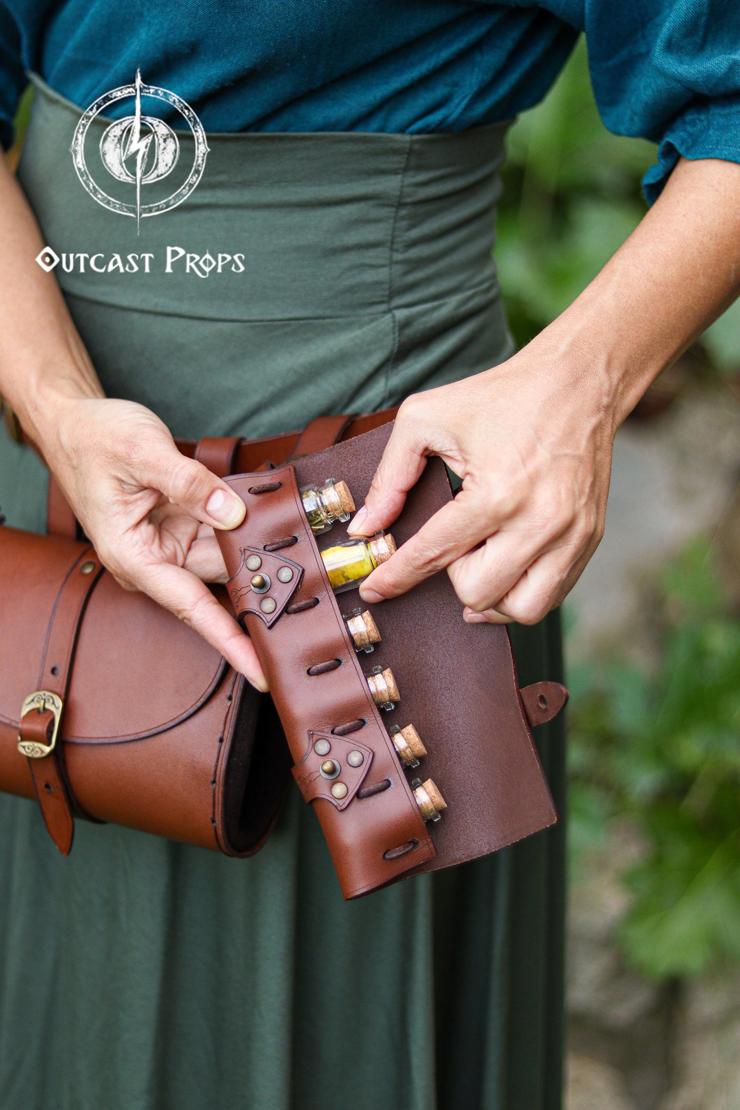 Close up of a brown leather potion case designed to hold six small corked vials, shown in the hands of a woman wearing a teal top and green skirt in a garden setting. She removes a yellow-filled bottle to demonstrate access and security. The handcrafted vegetable tanned leather holster features decorative rivets and tight loops that keep the bottles stable while moving. Ideal for alchemists, healers, cooks, wizards, or steampunk LARP characters who need a compact potion belt or spice holder.