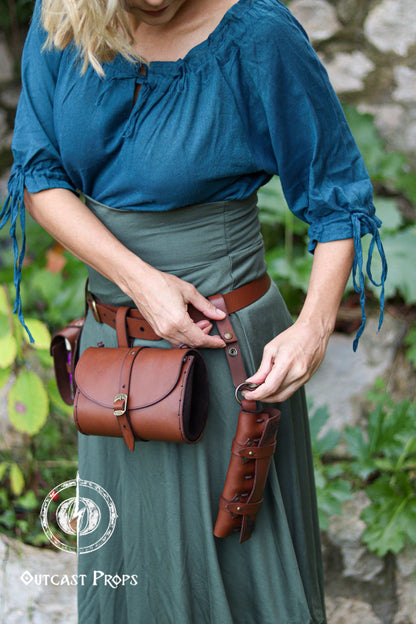 A woman in teal blouse and long green skirt adjusts the leather strap of a vertical potion holster hanging from her belt. The brown leather case holds six vials and sits neatly beside a matching round pouch, creating a functional alchemist belt kit. The scene shows how the potion holder attaches to the hip with snaps and rings, offering space-saving carry for LARP, steampunk, or fantasy costumes. Perfect for holding potions, herbs, spices, or props for roleplay characters.