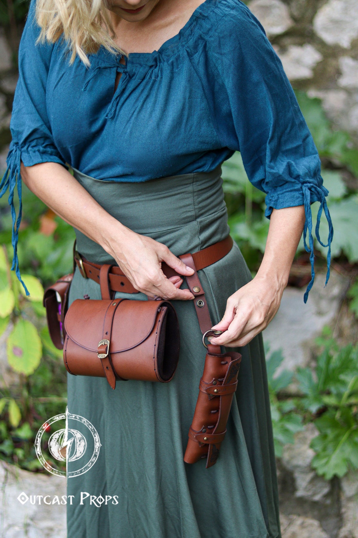 A woman in teal blouse and long green skirt adjusts the leather strap of a vertical potion holster hanging from her belt. The brown leather case holds six vials and sits neatly beside a matching round pouch, creating a functional alchemist belt kit. The scene shows how the potion holder attaches to the hip with snaps and rings, offering space-saving carry for LARP, steampunk, or fantasy costumes. Perfect for holding potions, herbs, spices, or props for roleplay characters.