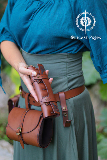 The woman holds the leather potion case detached from the belt to show its structure: a cylindrical brown leather body with embossed details, reinforced straps, and secure loops for six glass vials. She wears a teal top and green skirt, styled like an herbalist or wizard. The holster hangs from a wide belt and can be worn vertically for easy access. Ideal for LARP alchemists, potion makers, steampunk adventurers, healers, assassins or cooks carrying herbs or small items.