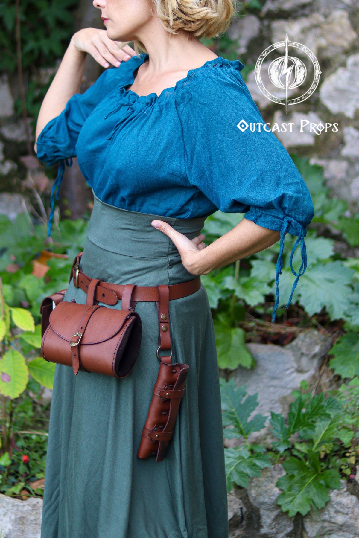 Side profile of a woman wearing a fantasy-inspired outfit with a wide green skirt and teal blouse, showing the leather clasp secured to her belt. A brown cylindrical potion holster and matching round pouch hang next to it, giving a clear sense of how multiple pieces can be attached. The clasp strap sits flat against the skirt and features two snap-closure positions. Ideal for LARP alchemists, rogues, healers or cosplayers who want to carry gear easily and keep their belt organized.