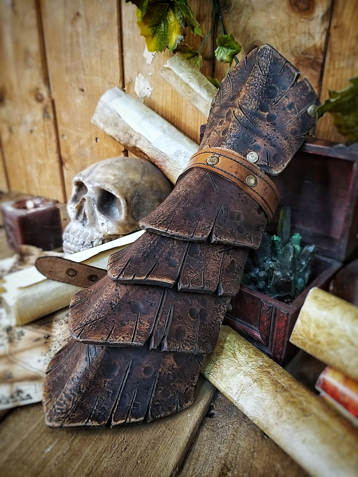 A brown handmade Dragon Scales Leather Bracer is worn on a man’s forearm, fully fastened and resting over a vintage map surrounded by fantasy props like a skull and candle. The layered leather plates resemble dragon hide, with an aged finish and rugged style. Perfect for LARP warriors, monster hunters or medieval cosplay. The leather is vegetable-tanned and hand-painted using Outcast Props’ signature weathering technique.