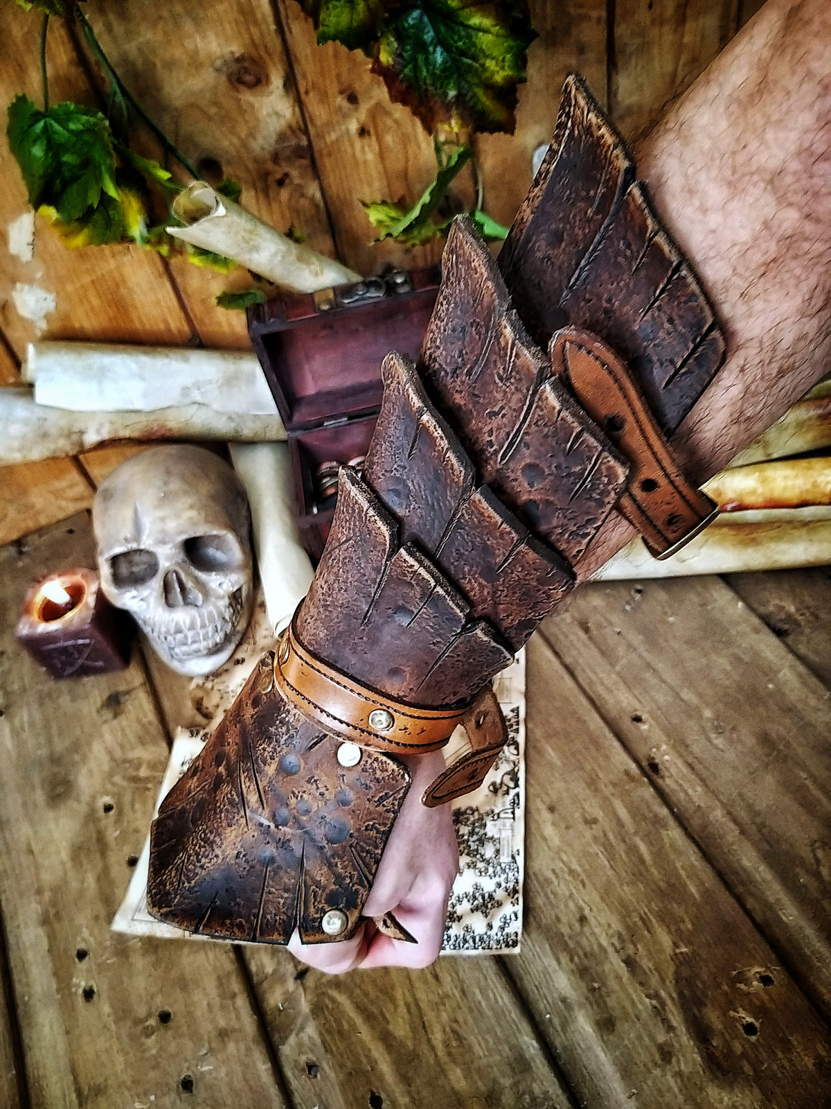 A brown handmade Dragon Scales Leather Bracer is worn on a man’s forearm, fully fastened and resting over a vintage map surrounded by fantasy props like a skull and candle. The layered leather plates resemble dragon hide, with an aged finish and rugged style. Perfect for LARP warriors, monster hunters or medieval cosplay. The leather is vegetable-tanned and hand-painted using Outcast Props’ signature weathering technique.
