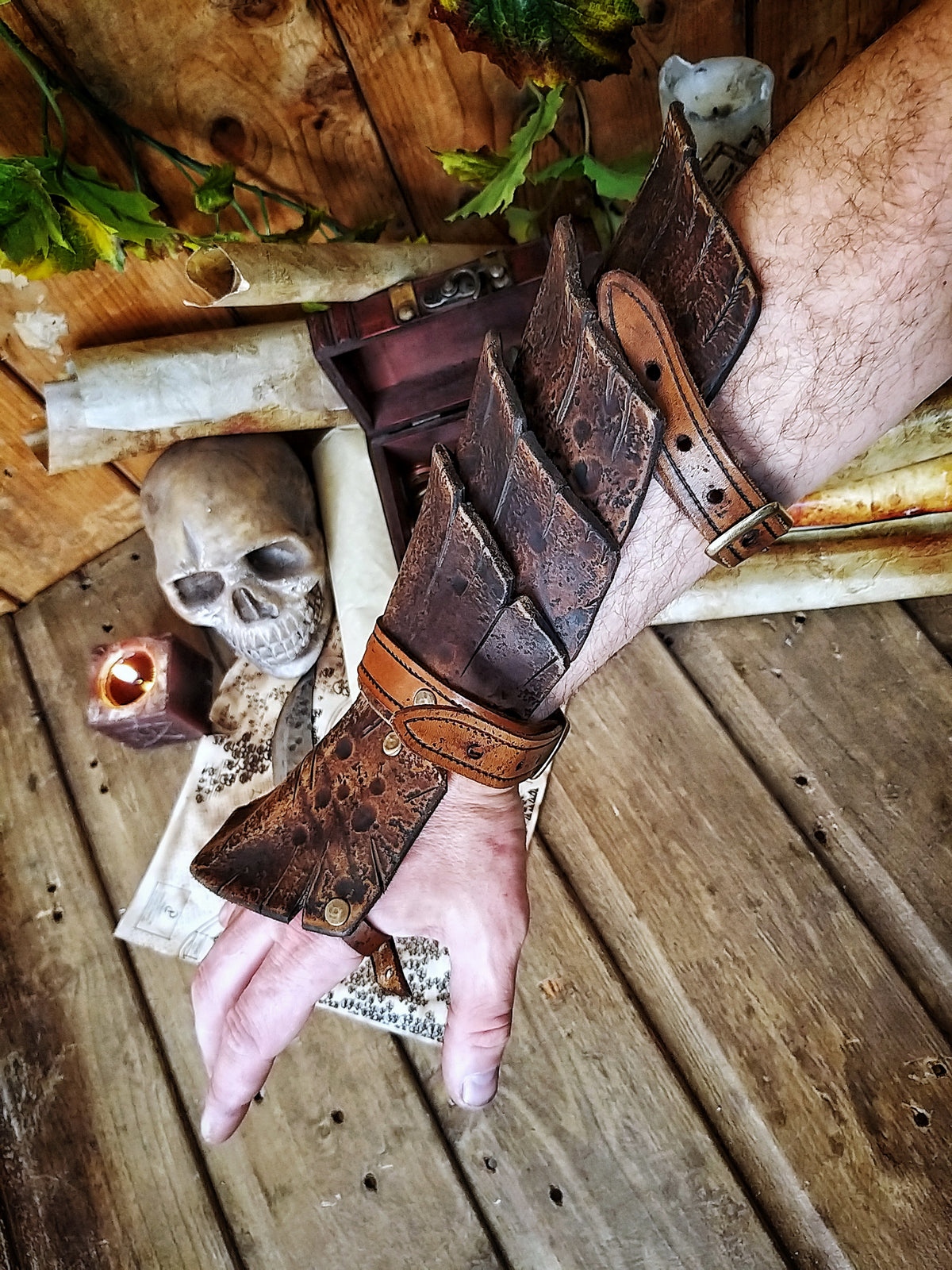 Close-up of a brown leather dragon scale bracer worn on a male hand, displayed over a vintage map with coins, a skull, and scrolls. This handmade bracer is ideal for LARP, pirate cosplay, monster hunters, or fantasy warriors. Crafted from premium vegetable-tanned leather and finished with Outcast Props' signature aging techniques, each piece is unique and tells a story straight from an epic adventure.