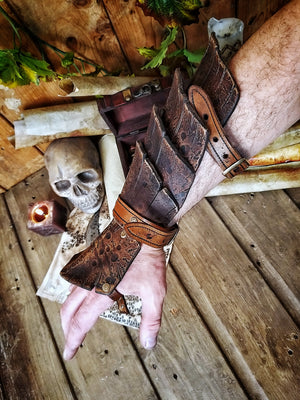 Close-up of a brown leather dragon scale bracer worn on a male hand, displayed over a vintage map with coins, a skull, and scrolls. This handmade bracer is ideal for LARP, pirate cosplay, monster hunters, or fantasy warriors. Crafted from premium vegetable-tanned leather and finished with Outcast Props' signature aging techniques, each piece is unique and tells a story straight from an epic adventure.