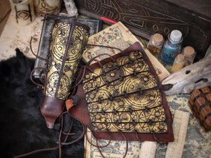 Golden fantasy bracers with matching armguard, engraved with magical runes and ritual symbols, shown in an arcane tabletop scene with scrolls and potions. Perfect for LARP witch armor, warlock costume or alchemist outfit. Handmade with aged leather and adjustable laces, these vambraces are a high-quality accessory for medieval, fantasy or occult cosplay enthusiasts.