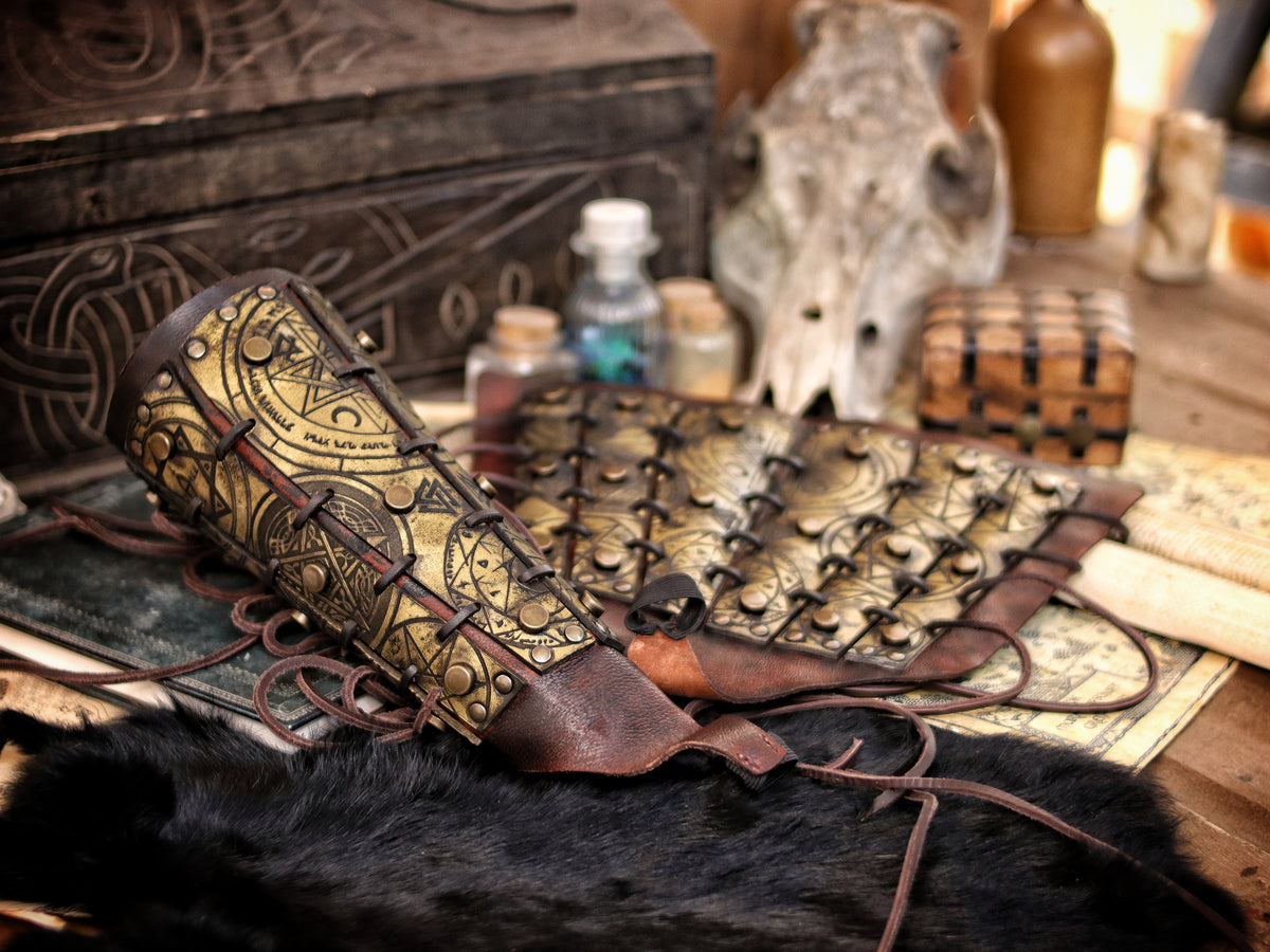 Golden fantasy bracers with matching armguard, engraved with magical runes and ritual symbols, shown in an arcane tabletop scene with scrolls and potions. Perfect for LARP witch armor, warlock costume or alchemist outfit. Handmade with aged leather and adjustable laces, these vambraces are a high-quality accessory for medieval, fantasy or occult cosplay enthusiasts.