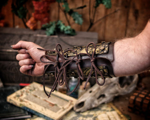 Golden fantasy bracers with matching armguard, engraved with magical runes and ritual symbols, shown in an arcane tabletop scene with scrolls and potions. Perfect for LARP witch armor, warlock costume or alchemist outfit. Handmade with aged leather and adjustable laces, these vambraces are a high-quality accessory for medieval, fantasy or occult cosplay enthusiasts.