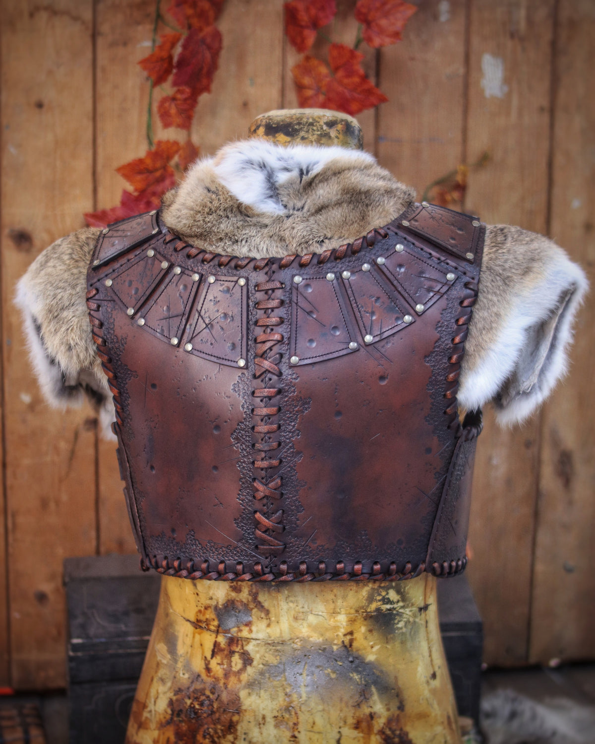 A handmade leather studded armor displayed on a wooden stand with a fur cape, featuring brown leather plates with studs, and a corded edge finish. Barbarian studded leather chest armor with rabbit fur cape attachment shown on mannequin. Perfect for fantasy LARP, Viking reenactment or cosplay, this full-grain leather armor is aged by hand and paired here with fur for an epic look. Adjustable buckle straps and visible stitching add to the rugged charm of this high-impact warrior costume piece.
