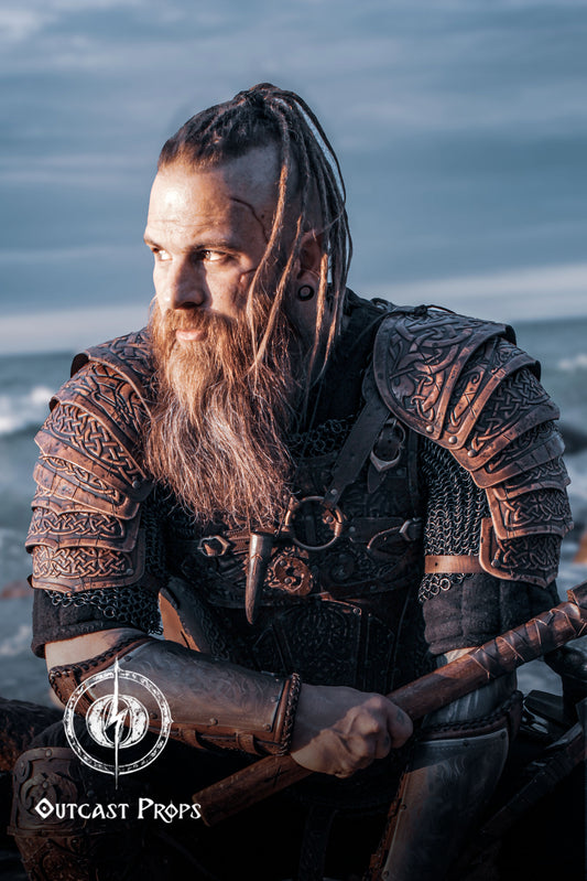 Bearded LARP warrior in full viking leather shoulder armor Sedrik, seated by the sea at sunset. The engraved Nordic pauldrons with layered leather plates feature detailed Celtic knots and fold for full mobility. Ideal for viking cosplay shoulder armor, Nordic warrior LARP gear, celtic leather pauldrons for fantasy costumes or medieval shoulder armor for immersive roleplay and conventions