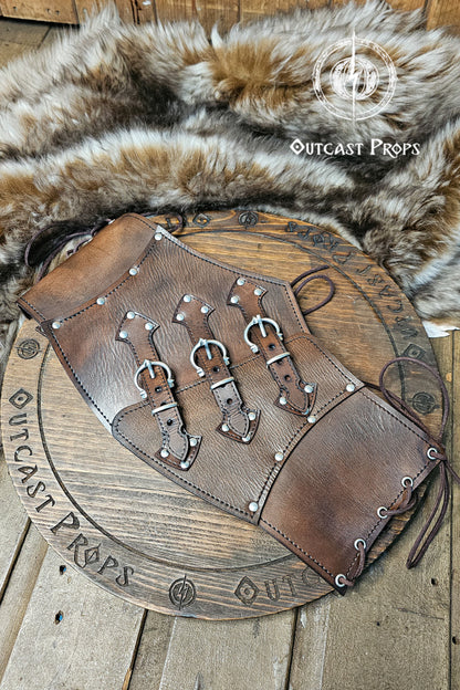 Handcrafted aged brown leather witch corset with nickel hardware displayed on a wooden board surrounded by fur. The waist cincher features triple buckle straps and side lacing, inspired by steampunk and gothic fashion. Ideal for ren faire or LARP costumes, this piece showcases Outcast Props’ signature aging technique and sustainable vegetable-tanned leather.