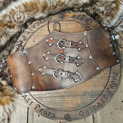 Handcrafted aged brown leather witch corset with nickel hardware displayed on a wooden board surrounded by fur. The waist cincher features triple buckle straps and side lacing, inspired by steampunk and gothic fashion. Ideal for ren faire or LARP costumes, this piece showcases Outcast Props’ signature aging technique and sustainable vegetable-tanned leather.