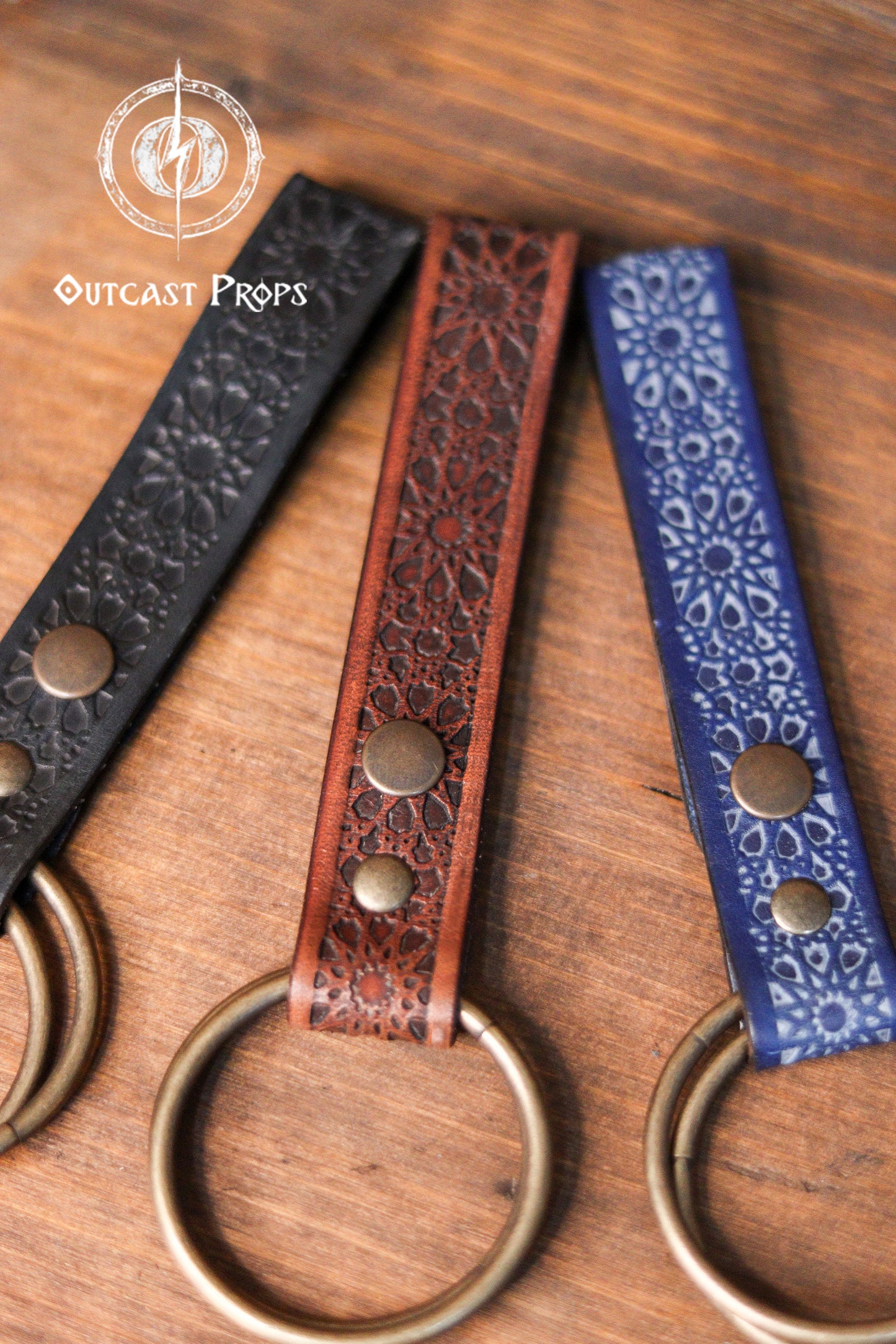 Close-up of three Persian leather skirt hikes in black, brown, and blue, displayed on a round wooden board engraved with Outcast Props branding. Each handcrafted leather skirt hike is embossed with Persian Alhambra-inspired patterns and features brass O-rings and snap buttons. This premium LARP and cosplay accessory adds functional flair to medieval, fantasy, and renaissance fair belts, perfect for enhancing any fantasy character’s gear.