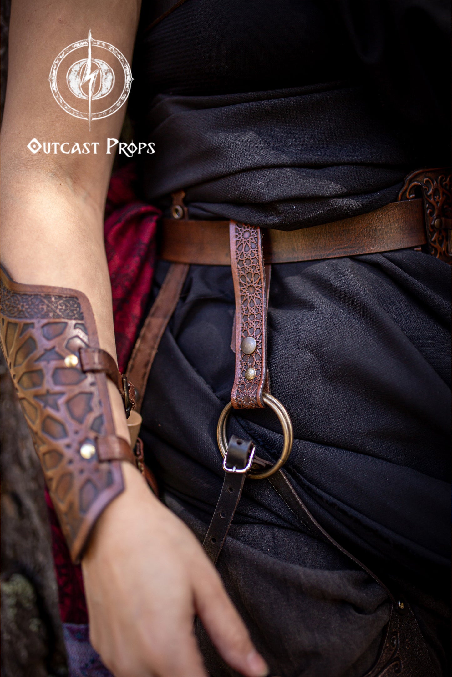 Medieval Persian leather skirt hike in brown, attached to a brown belt and black layered skirt on a fantasy LARP outfit. The accessory displays intricate Alhambra and Persian-inspired patterns and a brass O-ring. This handcrafted leather skirt hiker is designed for ren faire, LARP, fantasy costumes, and cosplay outfits, providing both a practical way to lift skirts or carry accessories and a beautiful decorative detail.