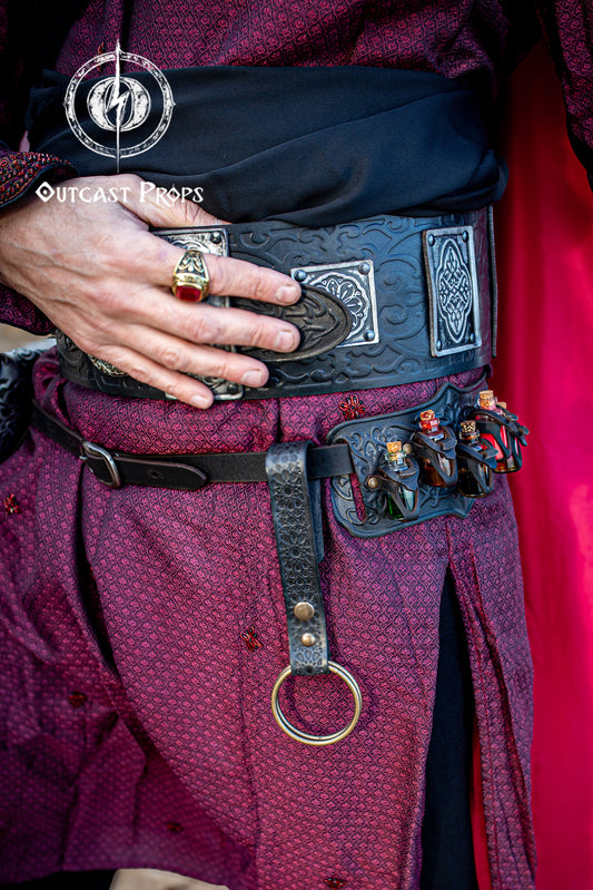 Black medieval Persian leather skirt hike with Alhambra-inspired patterns, worn on an ornate black and silver fantasy belt at a ren faire event. This LARP cosplay accessory features a large brass O-ring and aged snap button, used to hold fabric or suspend items. Perfect for adding practical elegance to renaissance fair costumes, medieval outfits, and fantasy LARP gear, crafted in premium Spanish leather.