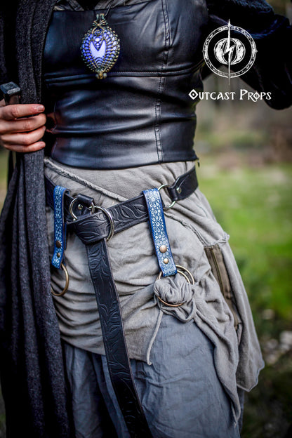 Two blue Persian leather skirt hikes attached to a black fantasy leather belt on a layered grey LARP costume. The skirt hikes, embossed with Arabic Alhambra motifs, feature brass O-rings and snap buttons for functional and decorative use. Ideal for medieval, ren faire, or fantasy cosplay outfits, these handcrafted leather accessories elevate your costume while offering practical versatility for LARP adventurers.