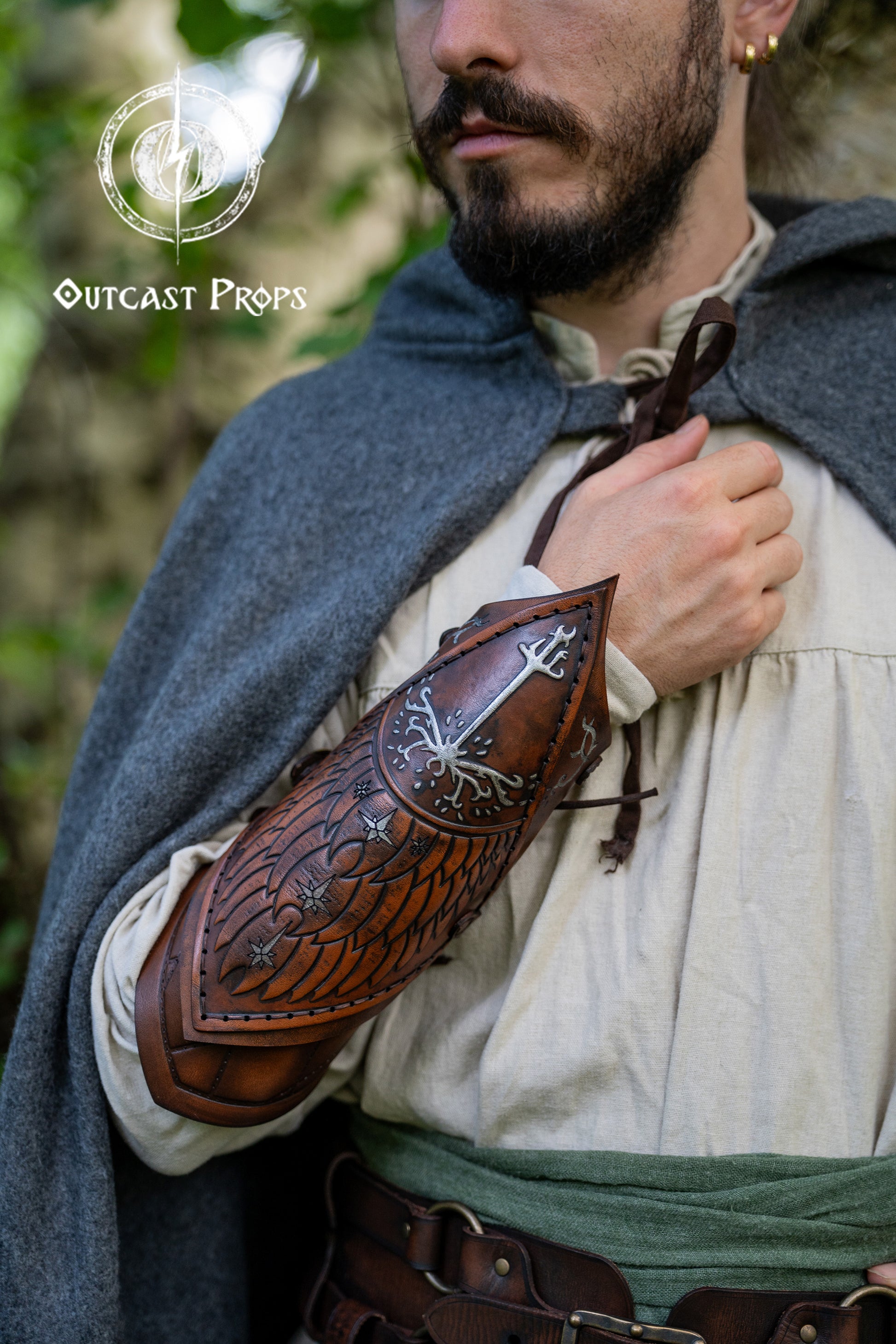 Fantasy ranger with brown embossed leather vambraces inspired by Gondor and the White Tree of LOTR. This handcrafted bracer set, worn over a medieval tunic and cloak, evokes Aragorn’s ranger gear. Perfect for Tolkien cosplay, LARP, reenactment or as a Gondorian warrior costume. Detailed elven engravings enhance the epic feel. Great for fans of Faramir, Boromir and the Citadel guards. Person wearing a detailed leather arm guard with Outcast Props logo. White tree leather bracers fantasy ranger armor LARP