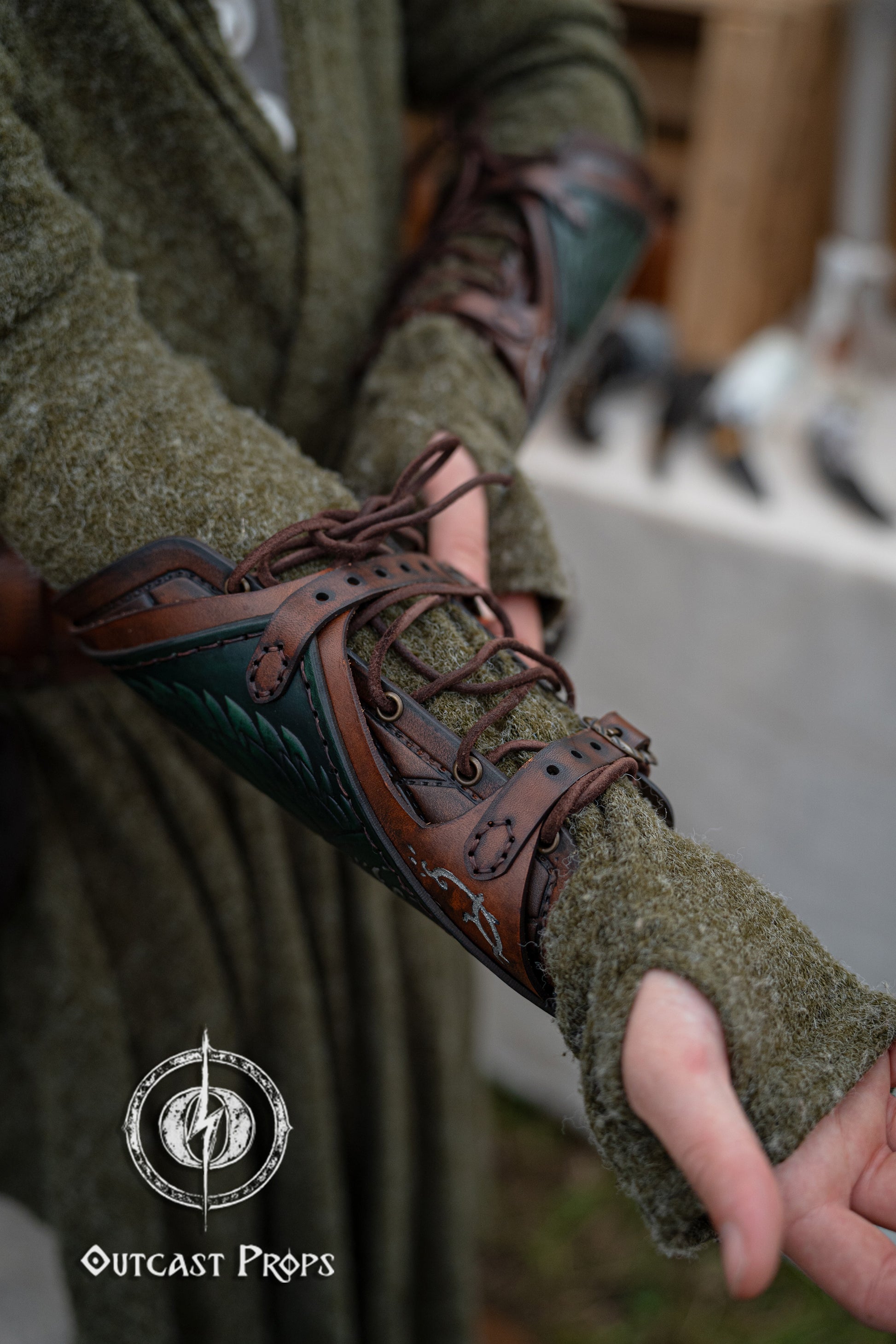 Close-up of green fingerless gloves with brown leather accents worn by a person, with 'Outcast Props' logo in the corner. Green Gondorian ranger vambrace shown from side angle, highlighting hand-laced leather and tree of Gondor emblem. Inspired by the warriors of LOTR like Faramir and Boromir, this bracer is crafted for LARP, cosplay, and medieval fantasy costumes. Worn over a wool tunic, it completes any elven ranger outfit or Tolkien-inspired reenactment. Perfect accessory for fantasy roleplay