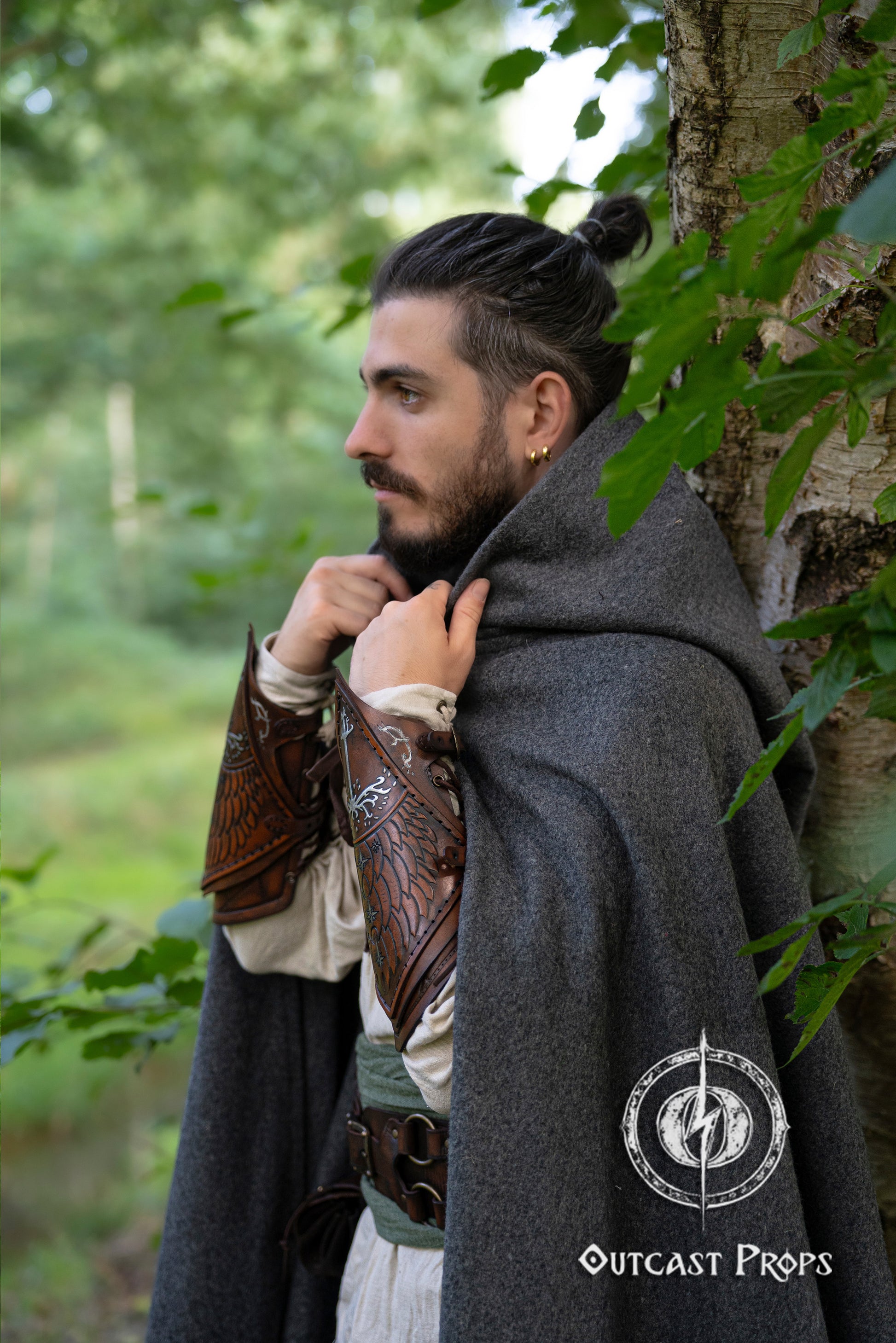 Man in a dark cloak with leather arm guards standing in a forest. Side profile of a fantasy ranger in a grey cloak, wearing brown Gondorian vambraces with White Tree embossing. Inspired by LOTR and Aragorn’s gear, these bracers are crafted for LARP, cosplay, and Tolkien fans. Elven feather engravings add authenticity. Ideal for medieval reenactments, roleplay, and portraying Gondor’s defenders or Citadel rangers. Premium handcrafted leather accessory.