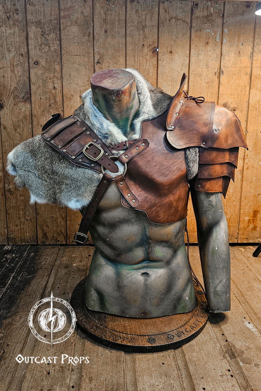 Leather barbarian armor set displayed on a muscular torso mannequin, featuring shoulder pauldrons, a chest harness, and thick grey rabbit fur mantle. Made from vegetable-tanned leather in aged brown tone, with weathered straps and metal buckles. Ideal for LARP, Viking cosplay or post-apocalyptic costumes. The fur and armor layers create a battle-worn barbarian look, full of texture and realism. Mannequin wearing a leather armor with fur accents against a wooden background, branded 'Outcast Props'.