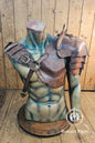 Mannequin bust wearing full barbarian leather shoulder armor set with crossed harness and riveted pauldrons. The armor is aged brown, handcrafted in vegetable-tanned leather with visible patina and hand-stitched details. Worn over a dark medieval shirt, this larp armor set includes sword scabbard on the back. Perfect for viking costume, post-apocalyptic larp, or warrior cosplay outfit.