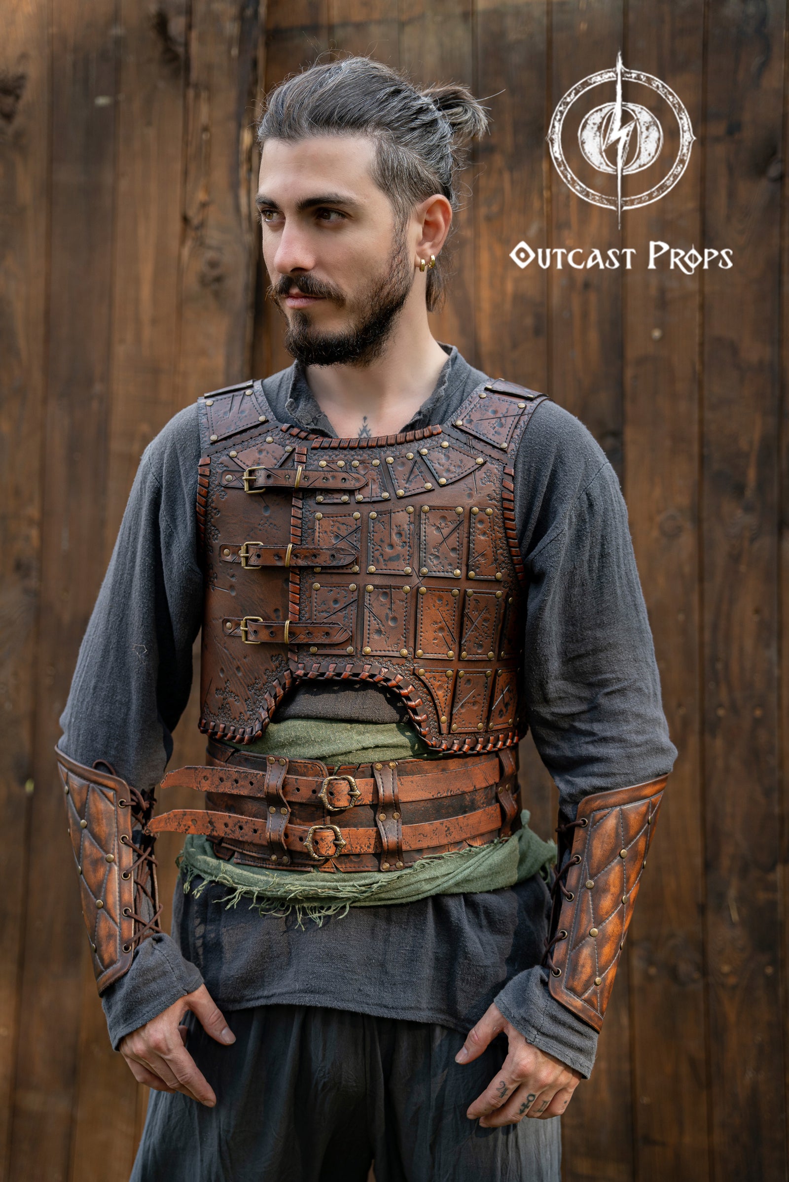 LARP leather armor set in dark brown tones with embossed battle scars and antique brass rivets. The warrior wears a full leather torso armor with triple buckle closure and matching leather vambraces. Designed for barbarian cosplay, fantasy roleplay and renfaire costumes, this handcrafted leather armor features Outcast Props' signature aging technique and embossed panels evoking a raw, tribal look. Viking Studded Leather Armor Chest Armor for Men LARP Armor Witcher Leather Armour Medieval Leather Armor 
