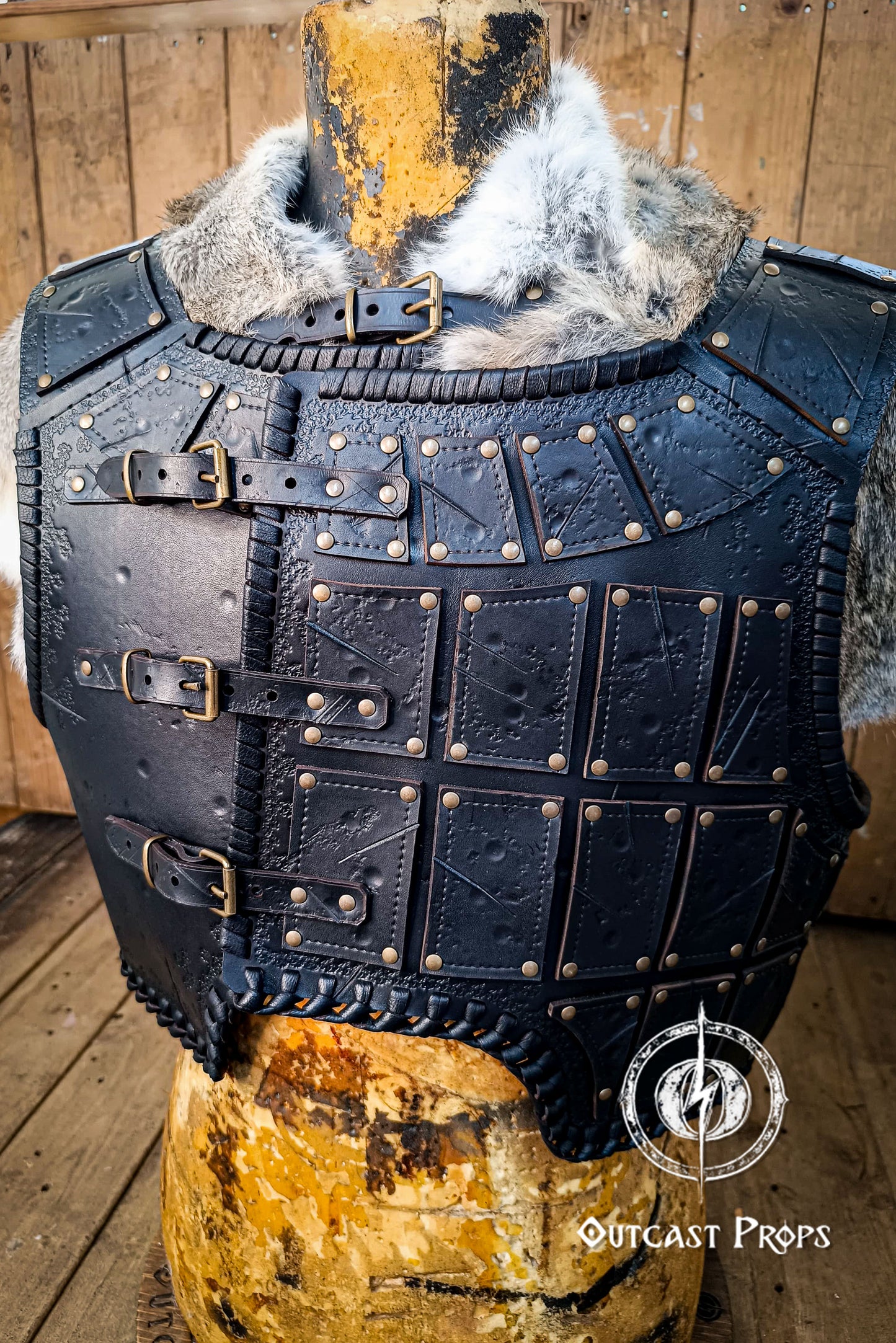Black leather armor vest with studs on a wooden mannequin, Outcast Props brand visible. A handmade leather studded armor displayed on a wooden stand with a fur cape, featuring brown leather plates with studs, and a corded edge finish. Barbarian studded leather chest armor with rabbit fur cape attachment shown on mannequin. Perfect for fantasy LARP, Viking reenactment or cosplay, this full-grain leather armor is aged by hand and paired here with fur for an epic look. Adjustable buckle straps and visible stit