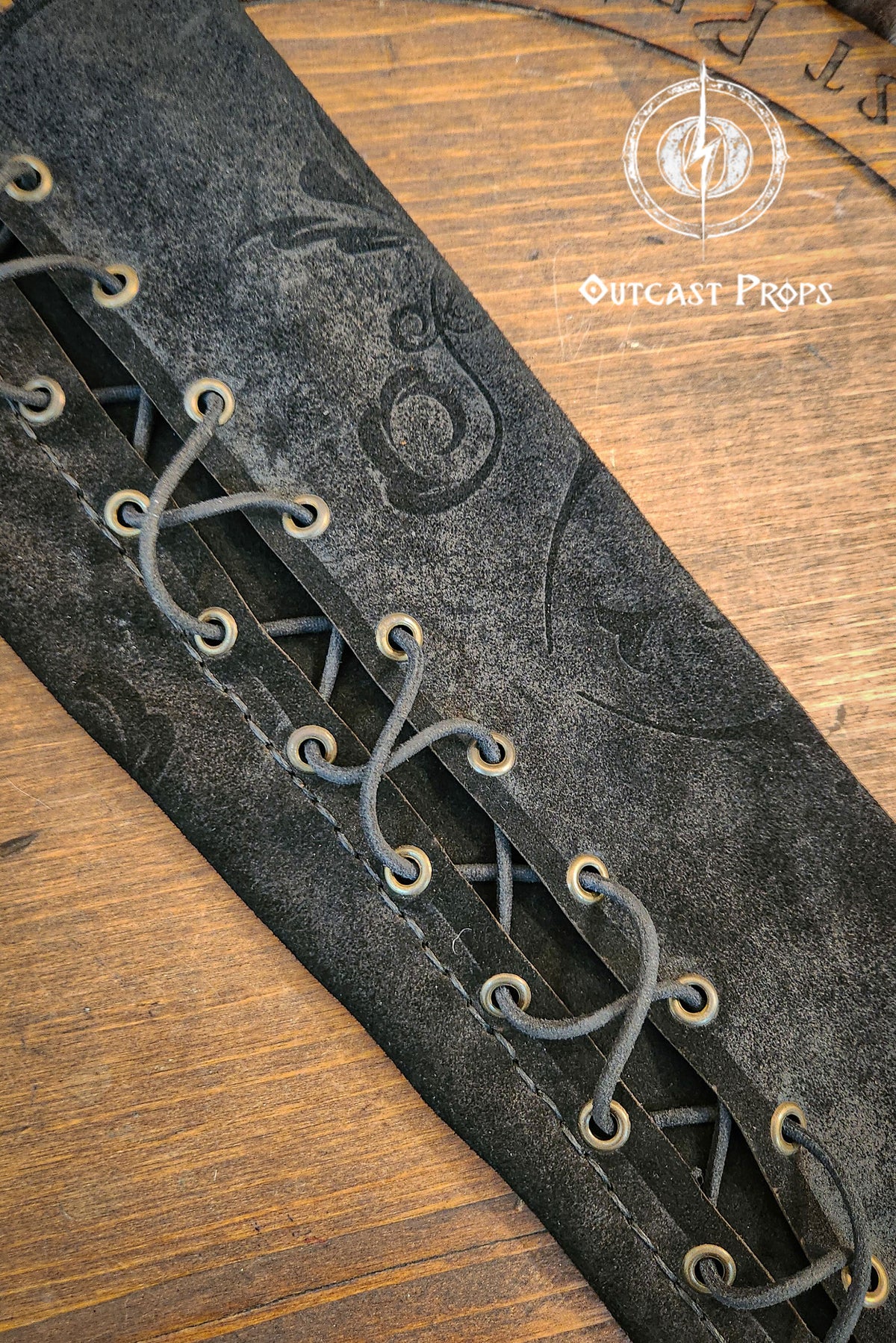 A close-up of a black leather arm guard with intricate details. The guard features a series of eyelets laced with a dark cord, creating a crisscross pattern. Embossed designs adorn the surface, and the Outcast Props logo is visible on a wooden background. A long black leather bracer sleeve rests on a round wooden board marked with runic style lettering and the Outcast Props emblem. Soft fur sits at the top of the frame, adding a noble and court inspired mood.