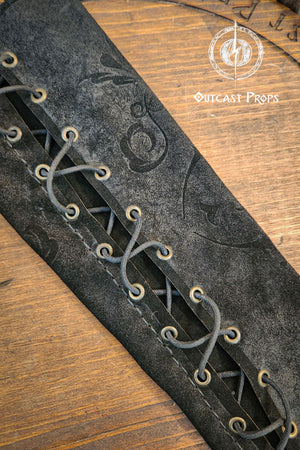 A close-up of a black leather arm guard with intricate details. The guard features a series of eyelets laced with a dark cord, creating a crisscross pattern. Embossed designs adorn the surface, and the Outcast Props logo is visible on a wooden background. A long black leather bracer sleeve rests on a round wooden board marked with runic style lettering and the Outcast Props emblem. Soft fur sits at the top of the frame, adding a noble and court inspired mood.