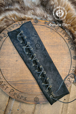 A black leather bracer sleeve with subtle floral embossing lies on a wooden table engraved with the Outcast Props logo, the suede surface catching soft light. The lacing runs in a neat crisscross through brass eyelets, giving it a court and noble aesthetic ideal for larp, renfaire and fae costume outfits. The close up shows the aged texture of the vegetable tanned leather, emphasizing artisan detail and elegant fantasy design. A black leather bracer with a lace-up design. Embossed patterns.
