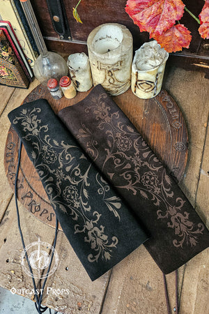 Two leather bracer sleeves, one black and one deep brown, lie side by side on a wooden table surrounded by candles, vials and antique books. Each sleeve shows detailed baroque floral embossing, giving them a noble court appearance perfect for larp, renfaire and fae costume characters. The scene evokes an alchemist or bard workspace, with warm light emphasizing the aged leather texture and artisan craftsmanship of the bracers.