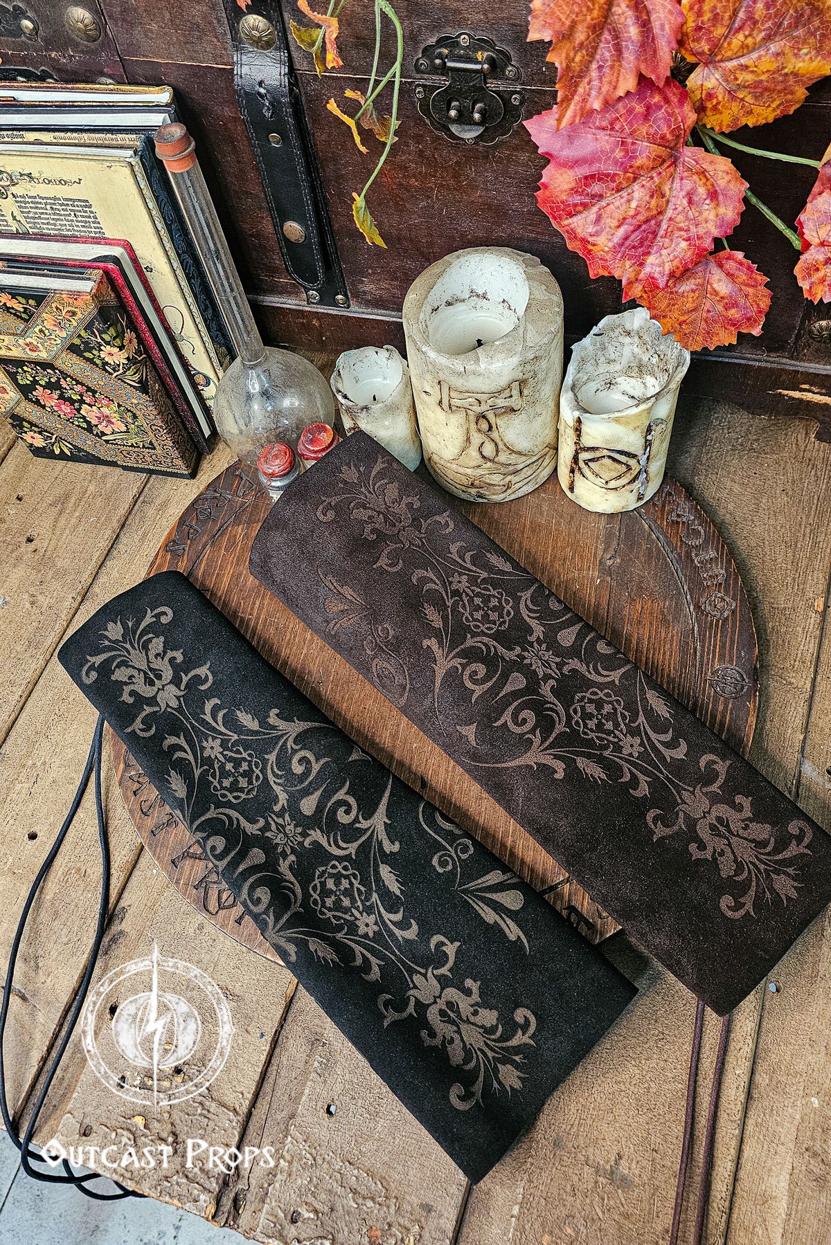 A pair of long embossed leather bracer sleeves rests on a wooden tabletop decorated with old candles, autumn leaves and vintage books. One sleeve is black and the other aged brown, both covered in ornate floral designs reminiscent of noble court fashion. The composition suggests a larp or renfaire dressing table, ideal for fae costume or fantasy characters. The lighting reveals the soft suede finish and detailed patterning on each bracer.