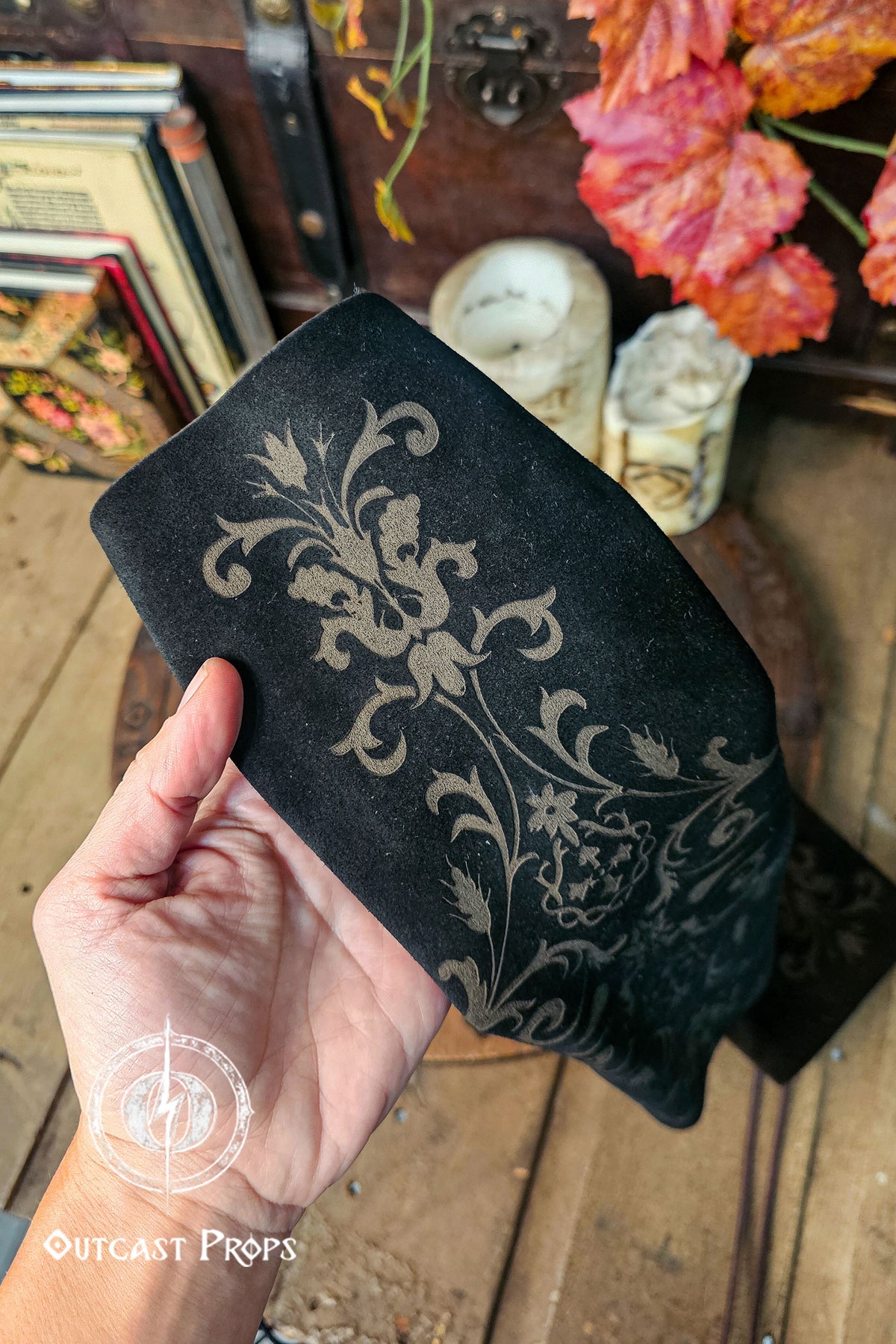 A close up of a hand holding a black leather bracer sleeve shows the embossed floral pattern in crisp detail. The suede surface bends slightly, revealing its softness and flexibility. Behind it sit bone-like candles, old books and warm autumn leaves, creating a noble court atmosphere suitable for larp, renfaire and fae costume outfits. The bracer’s floral motif and elongated shape highlight its role as an elegant fantasy leather sleeve.
