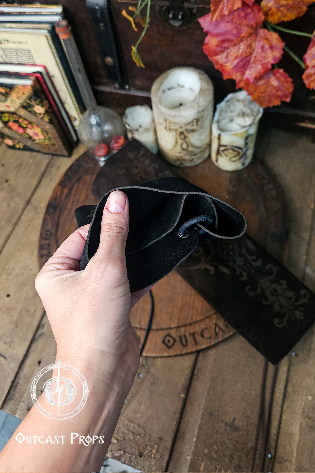 A hand gently squeezes the top of a long black leather bracer sleeve, showing the inner suede texture and flexibility of the material. Background props include antique style candles, books and a wooden board with the Outcast Props mark, creating a court and noble fantasy environment. The sleeve’s lacing channel is visible, hinting at its adjustable fit. Ideal for larp, renfaire or fae costume looks, the image emphasizes craftsmanship and durability.