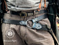 Person wearing a detailed belt with a large buckle, likely from a historical or fantasy costume, with 'Outcast Props' branding.  Waist-level shot of a leather wand holster belt styled with a long coat, ideal for dark wizard costumes. Great for Death Eater cosplay, wizard LARP, or gothic fantasy costumes. Combines functionality and style for Harry Potter-themed events and wizard duels. 