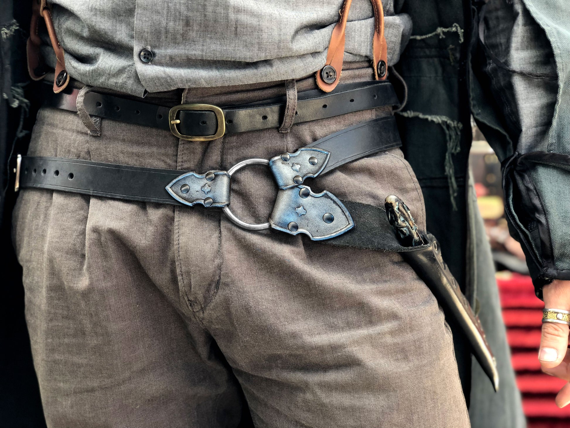 Waist-level shot of a leather wand holster belt styled with a long coat, ideal for dark wizard costumes. Great for Death Eater cosplay, wizard LARP, or gothic fantasy costumes. Combines functionality and style for Harry Potter-themed events and wizard duels. Person wearing a black leather harness on gray pants with a blurred background. Person wearing a detailed belt with a large buckle, gray pants, and a jacket.