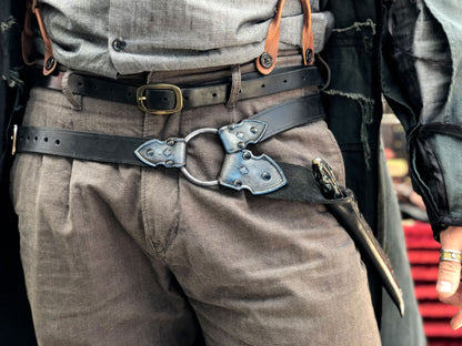 Waist-level shot of a leather wand holster belt styled with a long coat, ideal for dark wizard costumes. Great for Death Eater cosplay, wizard LARP, or gothic fantasy costumes. Combines functionality and style for Harry Potter-themed events and wizard duels. Person wearing a black leather harness on gray pants with a blurred background. Person wearing a detailed belt with a large buckle, gray pants, and a jacket.