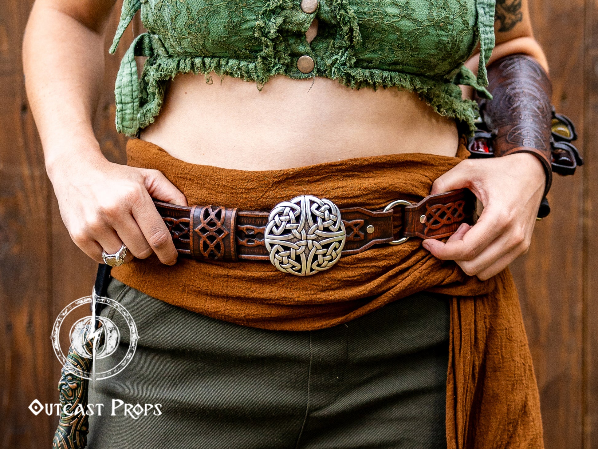Person wearing a green top and brown belt with a decorative buckle against a wooden background, with 'Outcast Props' logo. Celtic knot leather belt silver buckle larp belt for ren fair fantasy costume celtic belt unisex leather belt for renaissance outfit