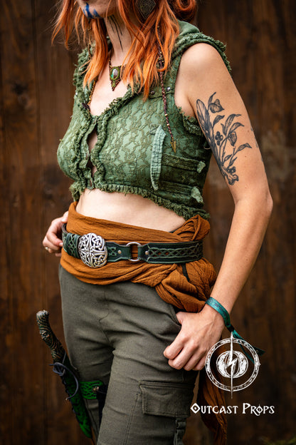 Person wearing a green textured top and brown pants with a tattoo on their arm, standing against a wooden background. Celtic knot leather belt silver buckle larp belt for ren fair fantasy costume celtic belt unisex leather belt for renaissance outfit