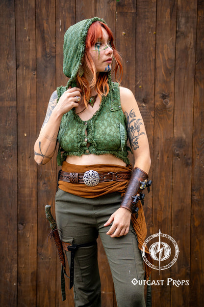 Person in a green hooded outfit with tattoos against a wooden background, Outcast Props logo visible. Celtic knot leather belt silver buckle larp belt for ren fair fantasy costume celtic belt unisex leather belt for renaissance outfit
