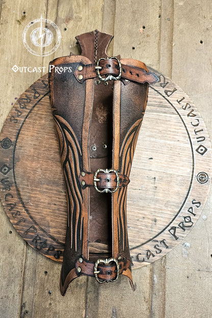 Aged brown Celtic leather greave handcrafted from full-grain vegetable-tanned leather with embossed flowing Nordic lines and three brass buckles. This medieval leg armor evokes Rohirrim horsemen and Viking warriors, combining authentic craftsmanship and durability for LARP and fantasy cosplay. The battle-worn finish and epic painting give each greave a heroic, cinematic look ideal for riders and explorers.